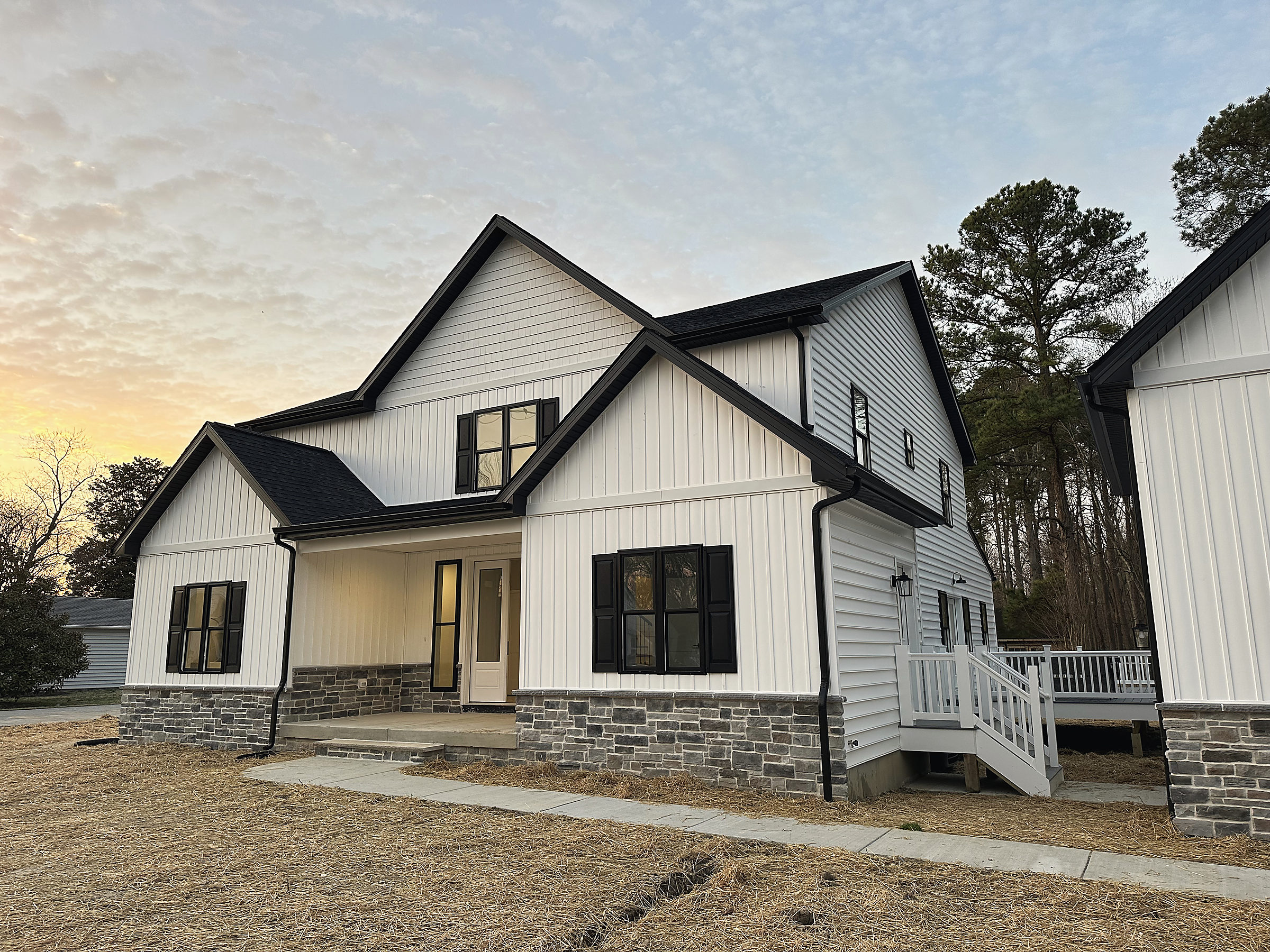 A white board-and-batten farmhouse with black windows and a stone skirt at dusk, built by Del Mar Construction in St. Michaels